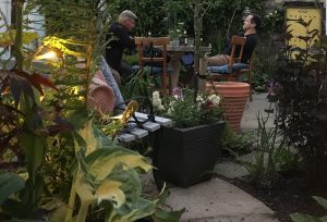 Two men relaxing at night in a warmly lit garden courtyard, sitting at a table surrounded by plants.
